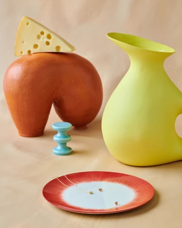 Still life shot of a triangular slice of cheese on an orange vase, near a yellow vase, a blue spool, and a white and red plate.