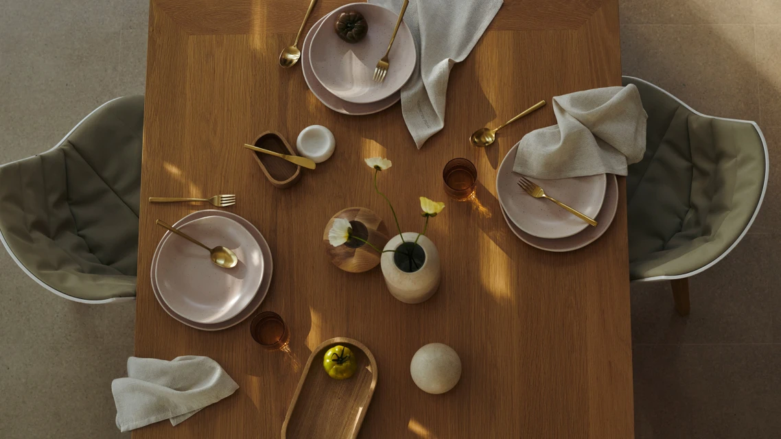 Overhead view of a wooden table set for dining with plates, gold cutlery, linen napkins, small flower vases, and contemporary chairs.