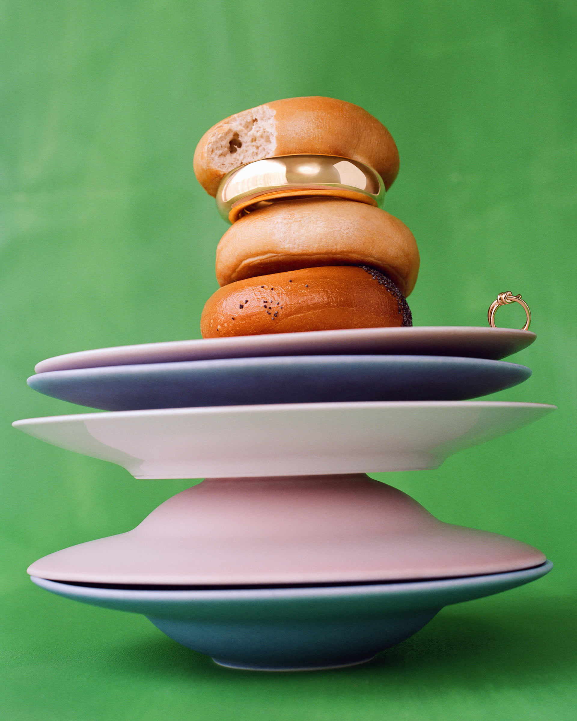Stack of bagels and jewelry piled on assorted plates on a green background for a graphic studio food and product shot.