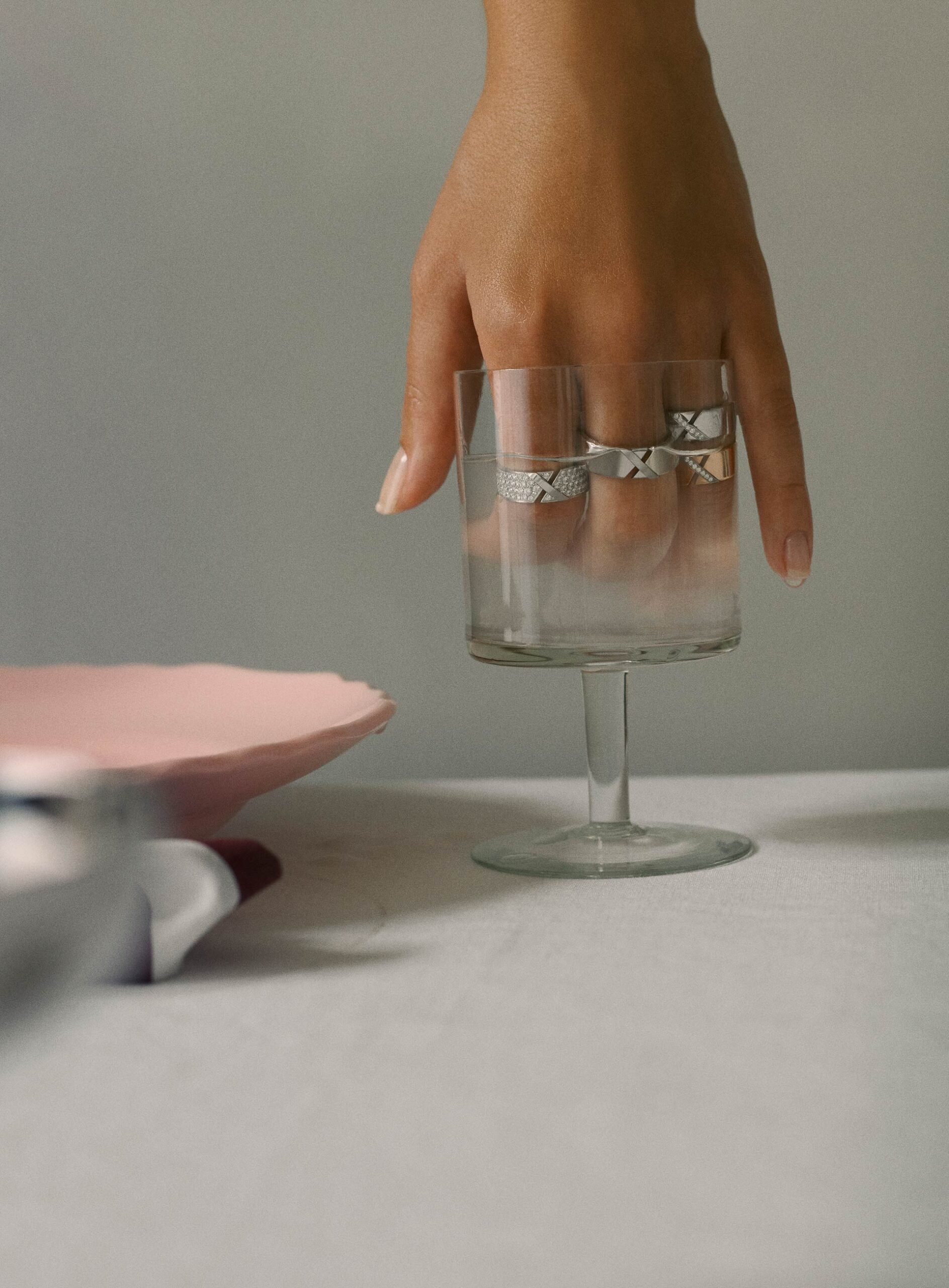Hand displaying rings on fingers, posed in a clear glass. Tabletop still-life showcases modern jewelry in a minimalist setting.