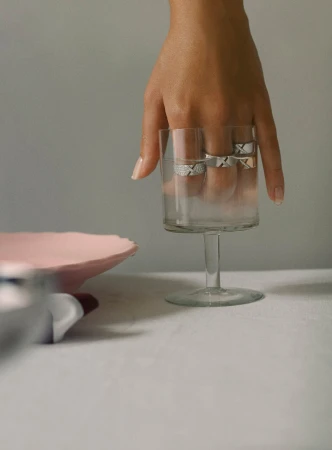 Hand displaying rings on fingers, posed in a clear glass. Tabletop still-life showcases modern jewelry in a minimalist setting.