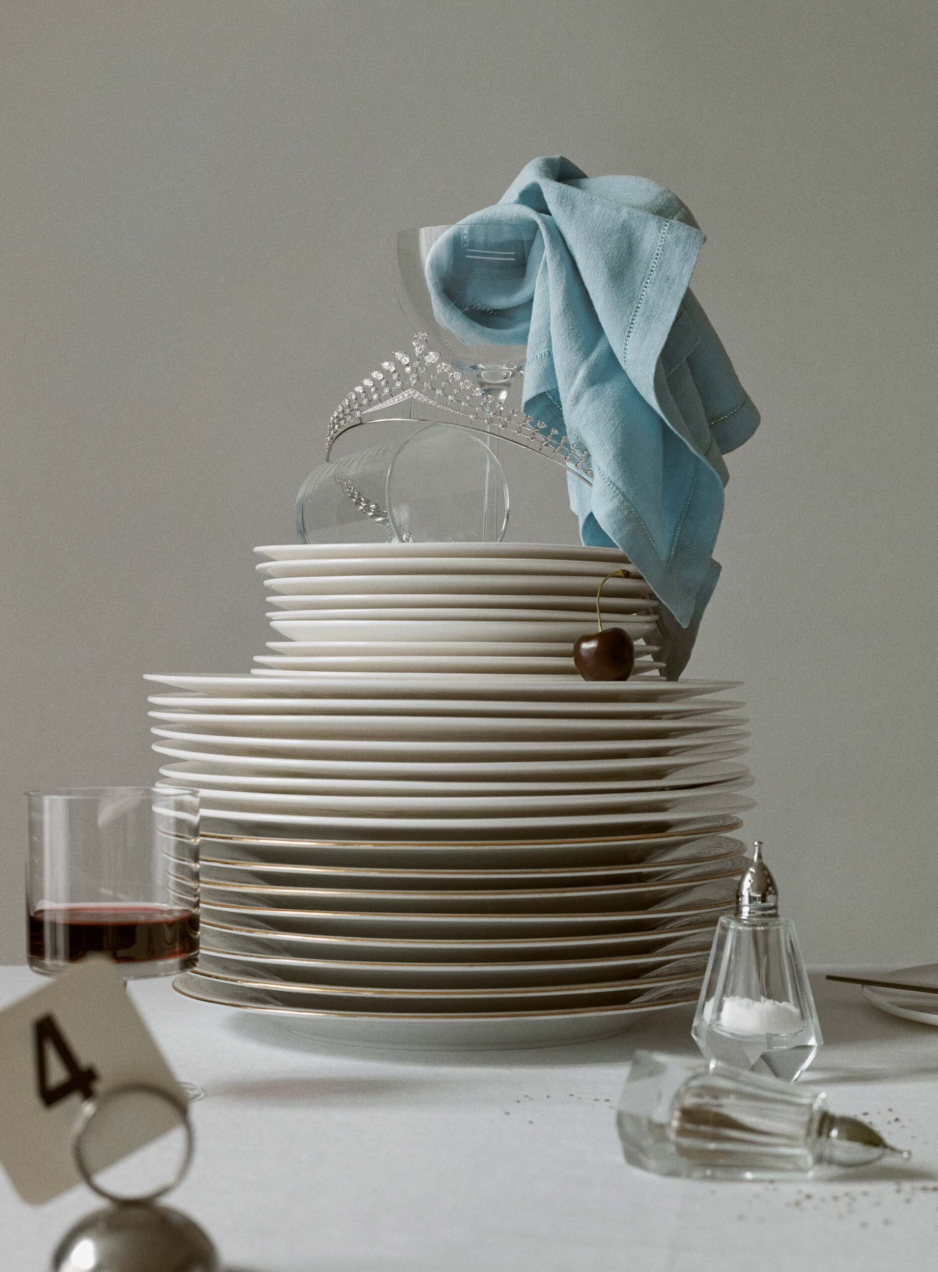 Still life of stacked plates, glassware, silverware, a tiara, and a cloth napkin, set on a table against a neutral background.