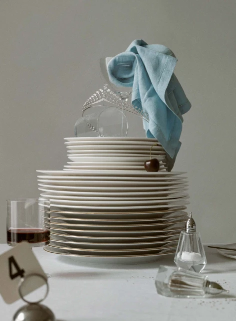 Still life of stacked plates, glassware, silverware, a tiara, and a cloth napkin, set on a table against a neutral background.