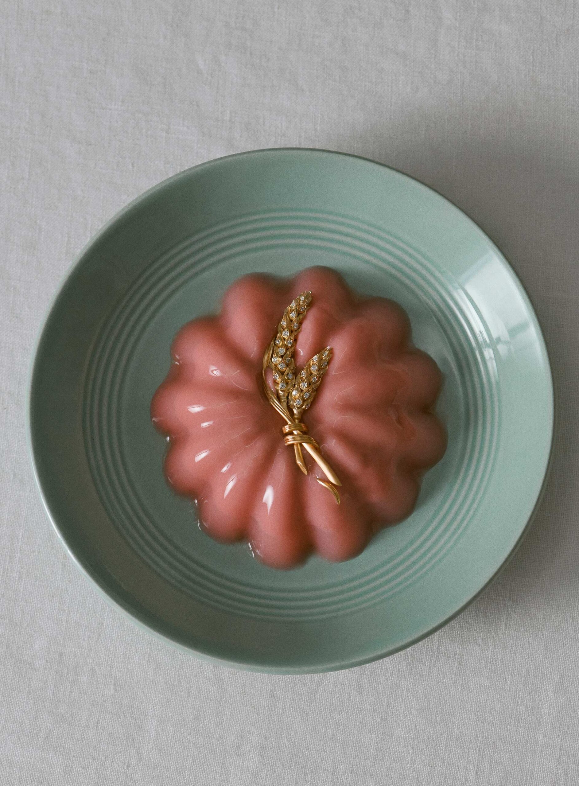 Pink jelly dessert on a teal plate with golden wheat stalk garnish, set against a neutral tablecloth backdrop, close-up studio shot.