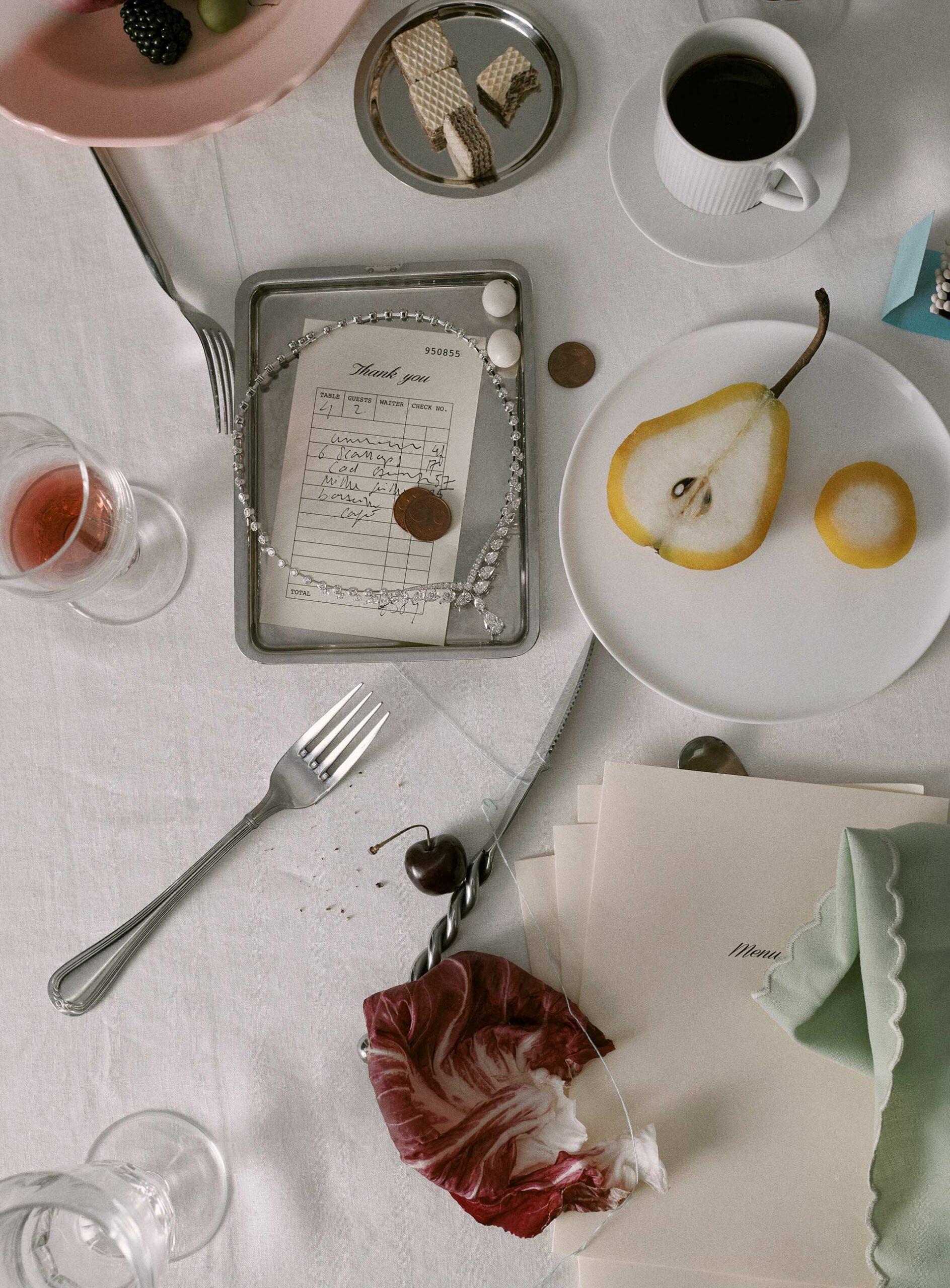 Overhead still life photo of a halved pear, a receipt with a diamond necklace, wafers, drinkware, flatware, and stationary on a white tablecloth.