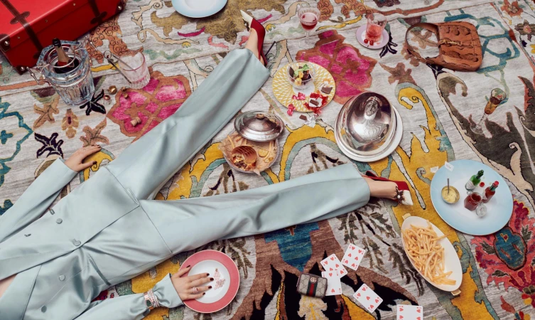 Overhead view of a person lying on a colourful patterned rug surrounded by plates of food, drinks, playing cards, and a red suitcase.