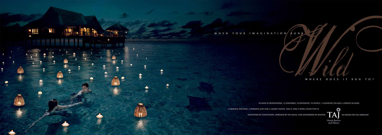 Evening shot of two people swimming in calm water with floating lanterns, against backdrop of a Taj resort on stilts.