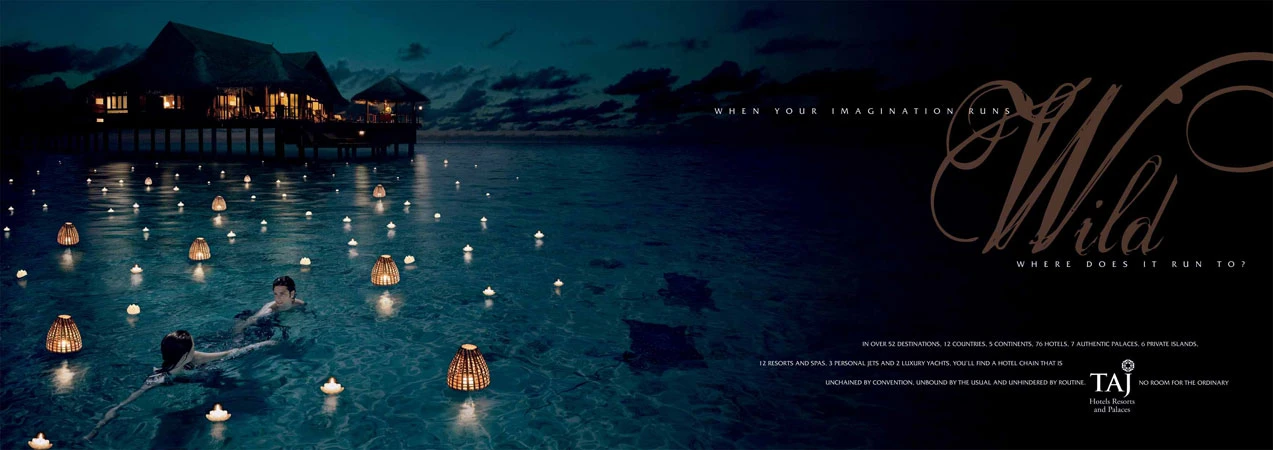 Evening shot of two people swimming in calm water with floating lanterns, against backdrop of a Taj resort on stilts.