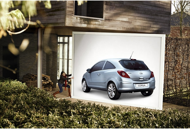 Silver Opel Corsa car on a large promotional board is positioned outside a modern house, with a woman standing nearby pushing it.