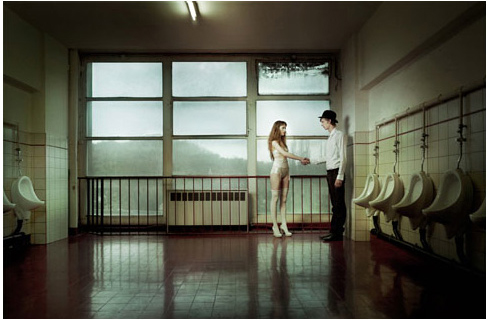 Man and woman in formal wear shake hands in an empty restroom with urinals, illuminated by fluorescent lighting.