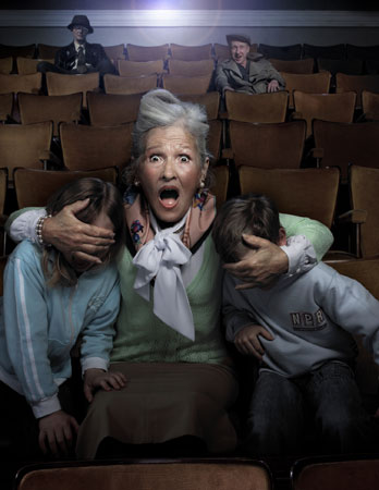 Older woman with an open mouth and startled expression is sitting in a movie theatre with two children, covering their eyes from the screen.