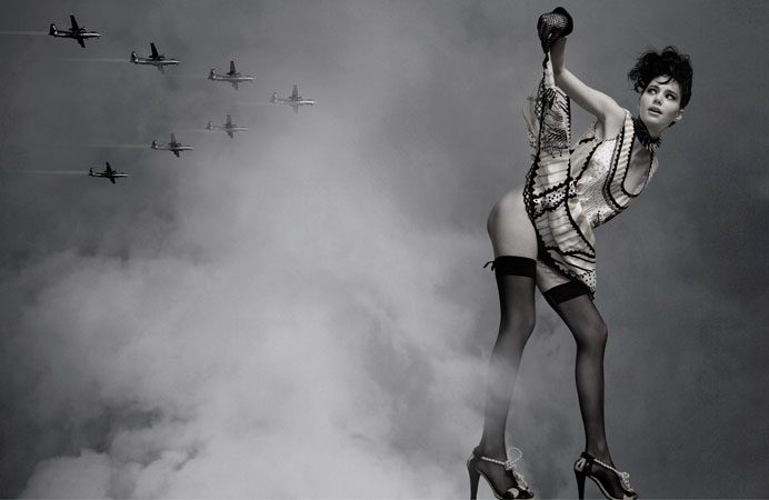 Black and white fashion shot of a woman in short dress, stockings and heels holding a glove with airplanes in the smoky background.