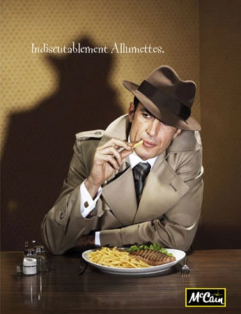Retro-style advertising photo of a man in a tan trench coat and fedora at a table eating french fries, with a dark shadow behind him.