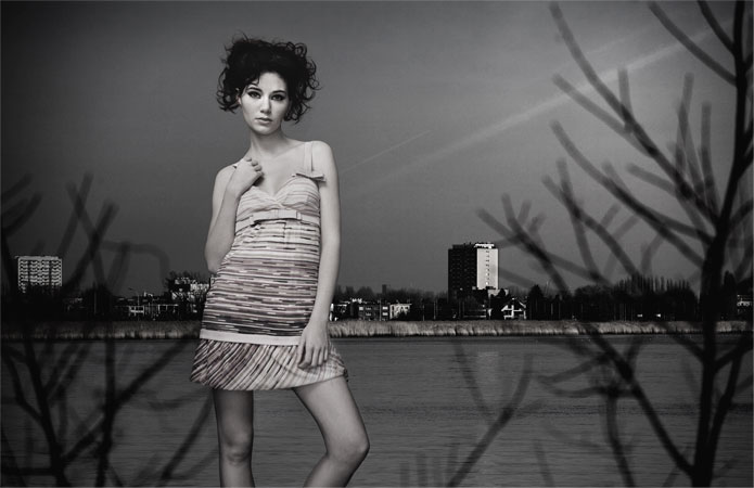 Adult woman in a dress poses outdoors near a body of water with buildings in the background, in a black and white fashion-style shot.