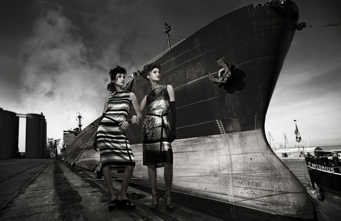 Two women in dresses pose in a monochromatic fashion shot beside a large ship with a cloudy sky, creating a dramatic and vintage aesthetic.