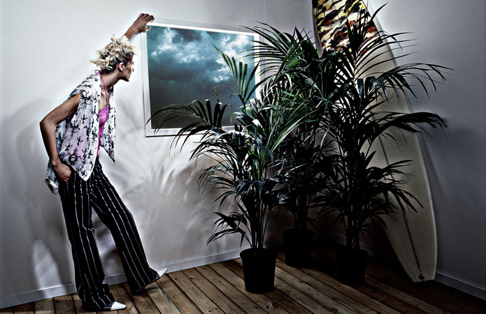 Woman in striped pants admires a framed storm cloud print in a minimal interior with potted palms and a surfboard.