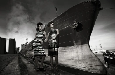 Two female models posing on a dock in front of a massive ship hull, photographed by Kurt Stallaert.