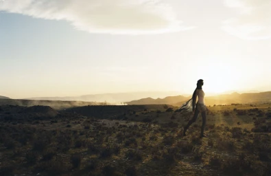 Silhouette of a woman walking across a rugged, arid landscape at sunset with a flowing scarf trailing behind her