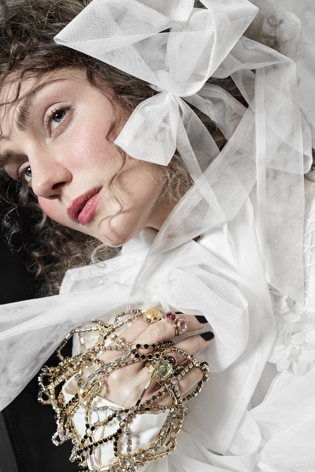 Portrait of a woman with fair skin, wearing a white ruffled blouse and bow, holding strands of gold-toned beaded jewelry indoors.