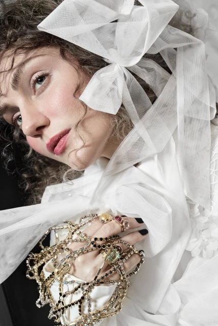 Portrait of a woman with fair skin, wearing a white ruffled blouse and bow, holding strands of gold-toned beaded jewelry indoors.