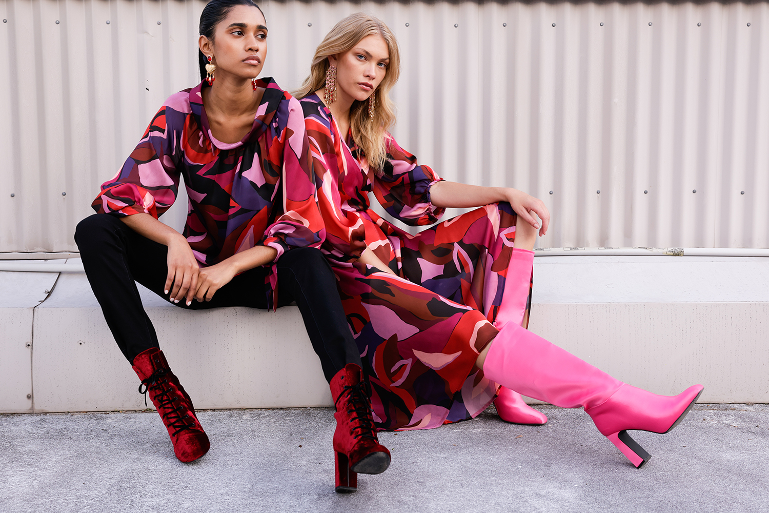 Two fashion models in colorful print tops, dark pants, and high-heeled boots pose outdoors against a textured background in a medium shot.