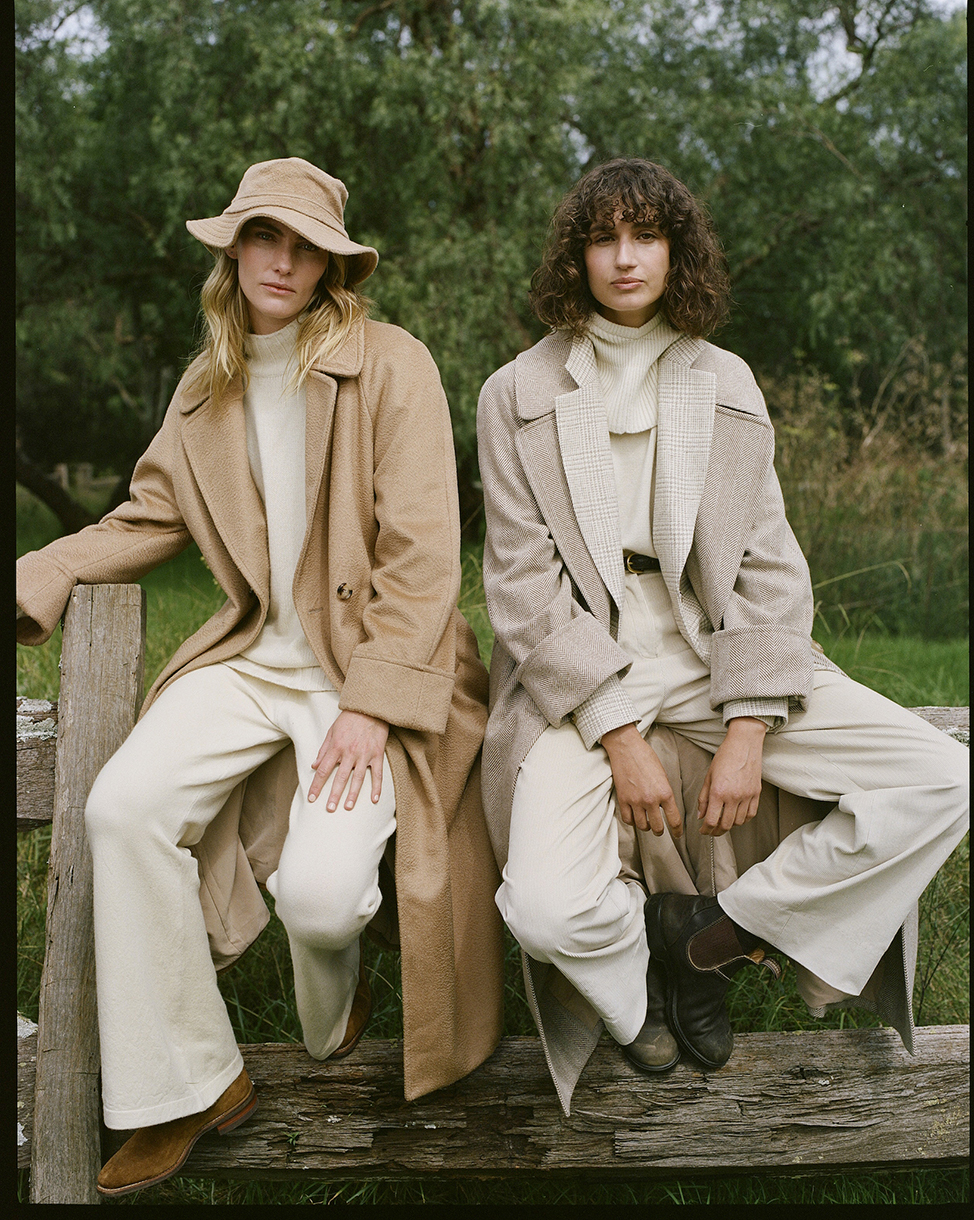 Two women in neutral-toned coats and pants sit on a wooden fence in a rural setting, exuding a relaxed, earthy fashion vibe.