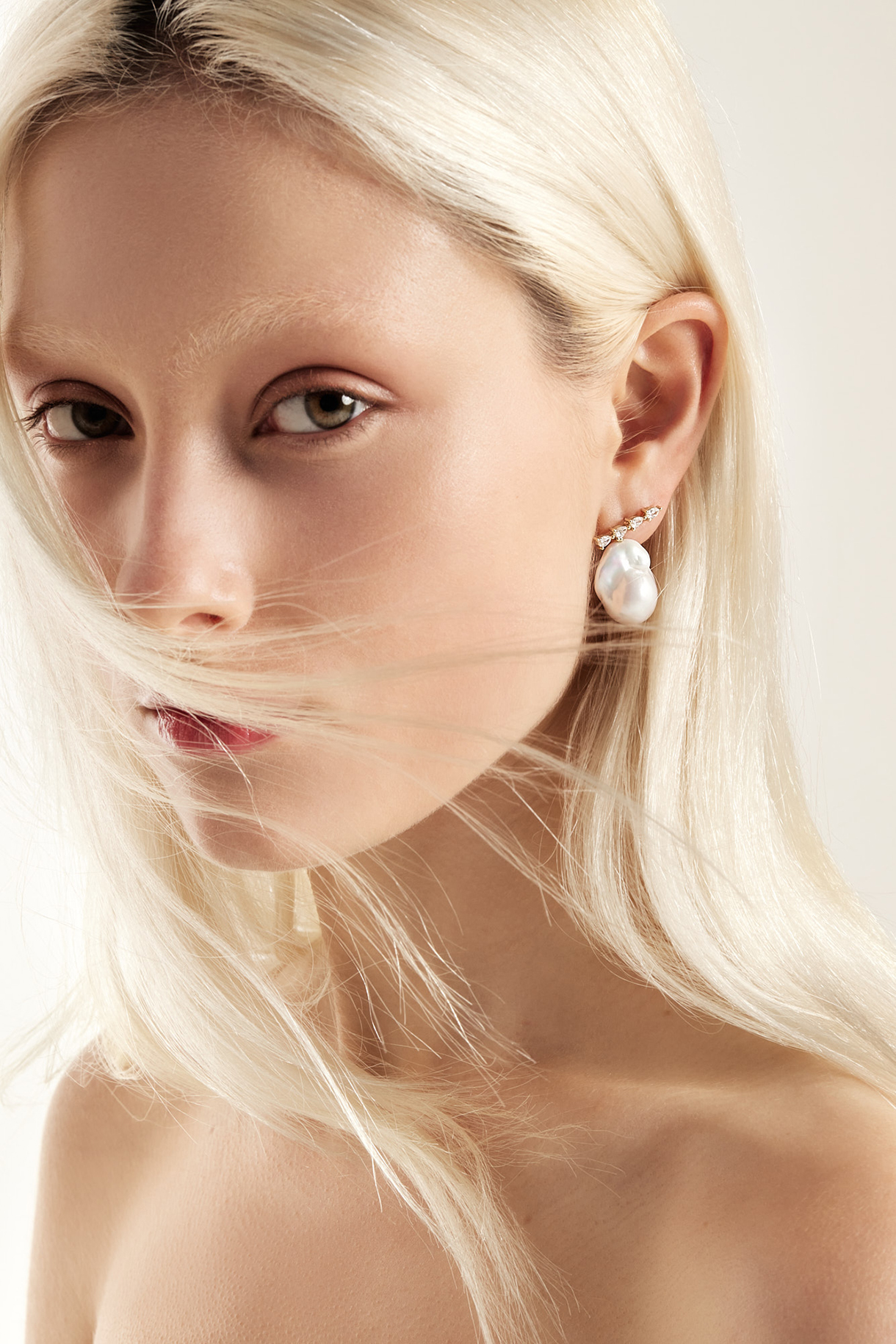 Close-up studio portrait of a woman with fair skin, light blonde hair covering half her face, and prominent baroque pearl earring.