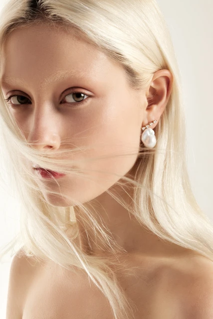 Close-up studio portrait of a woman with fair skin, light blonde hair covering half her face, and prominent baroque pearl earring.