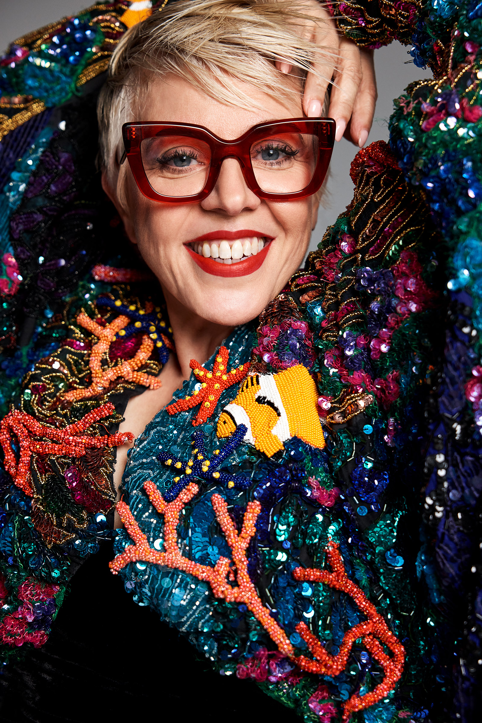 Close-up portrait of smiling woman wearing glasses and a vibrant beaded jacket with coral and clownfish designs against a grey backdrop.