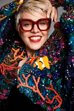 Close-up portrait of smiling woman wearing glasses and a vibrant beaded jacket with coral and clownfish designs against a grey backdrop.