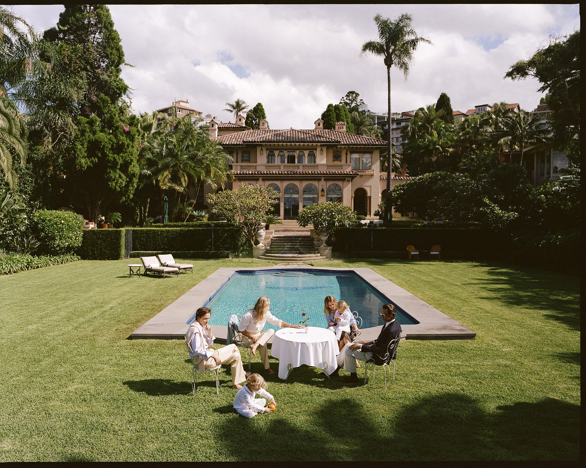 Family of five, including three children, seated outdoors around a white-clothed table on green lawn in front of swimming pool and estate.