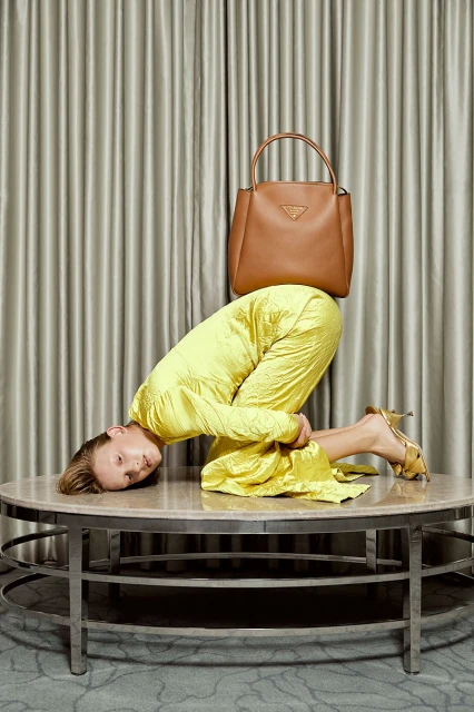 Woman in yellow crouches on table beneath Prada bag, fashion-forward shot, against curtain backdrop in a studio environment.