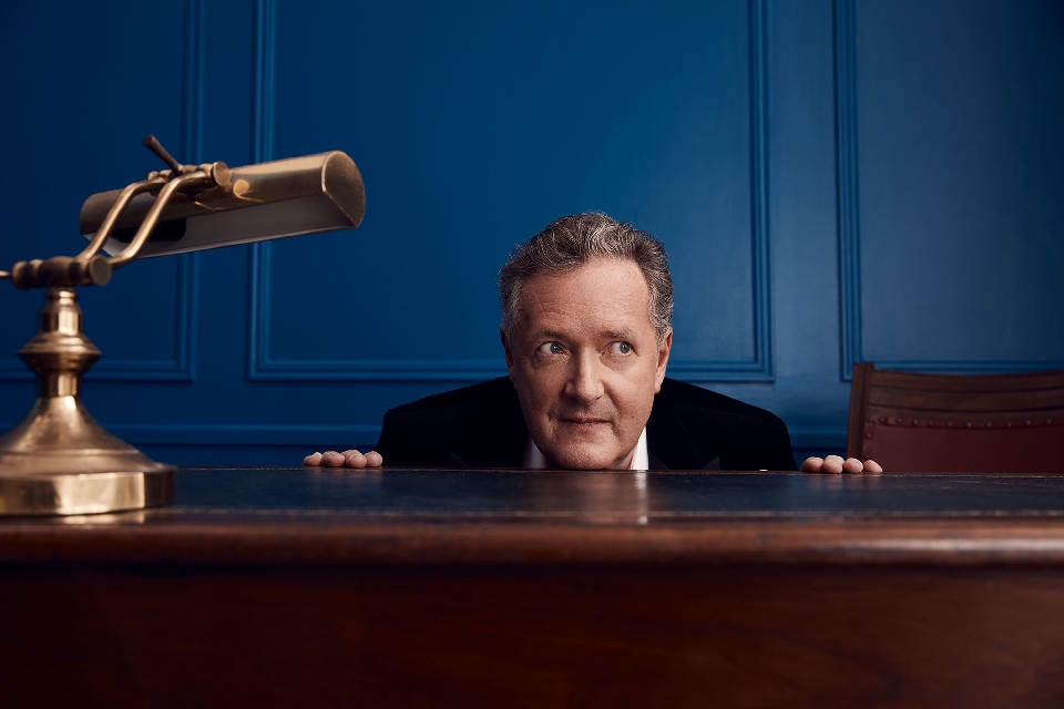 Portrait of a man in a dark suit behind a wooden desk with a brass lamp, set against a classic blue paneled wall background.