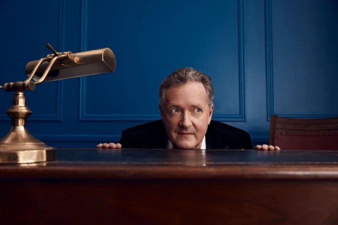 Portrait of a man in a dark suit behind a wooden desk with a brass lamp, set against a classic blue paneled wall background.