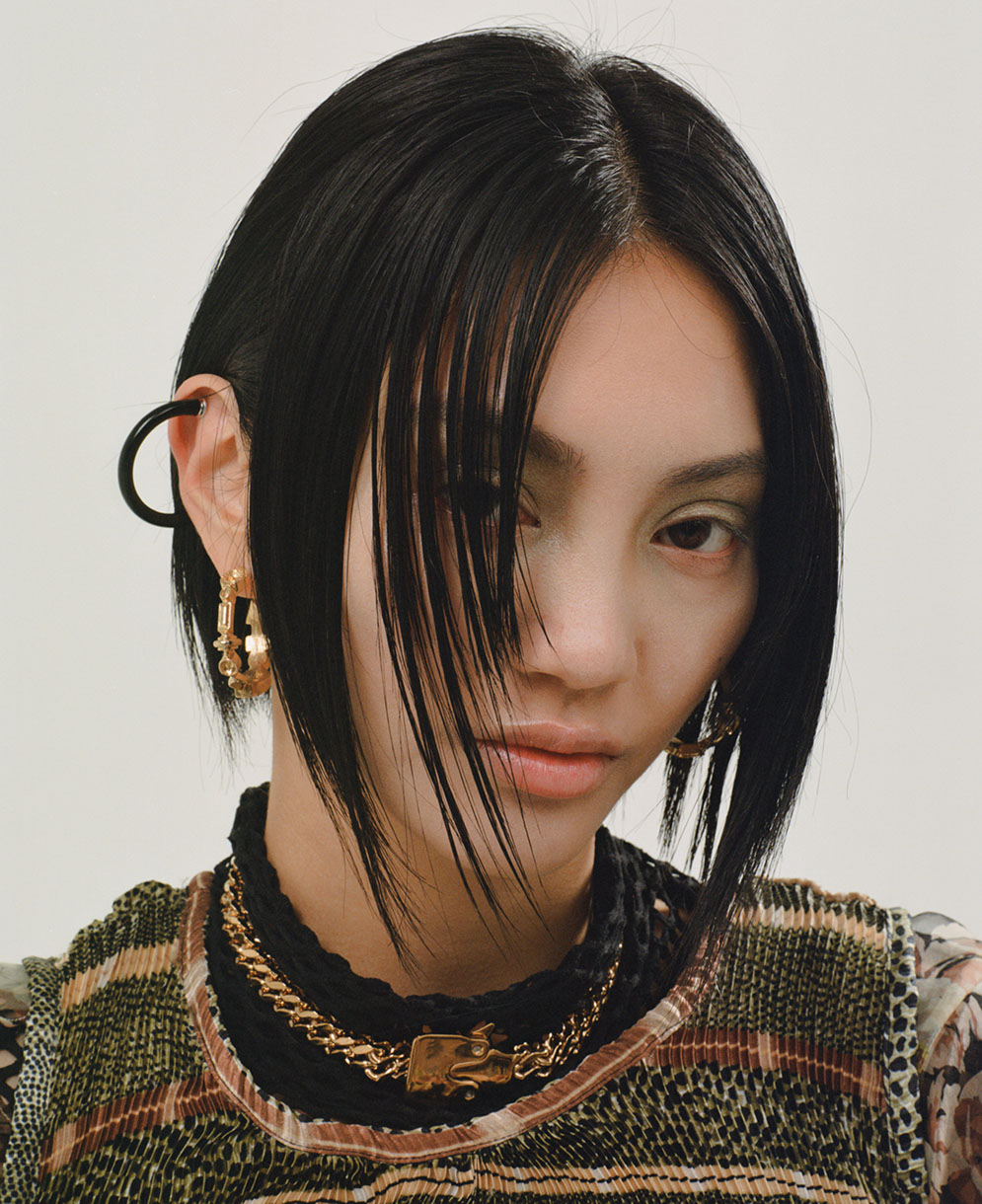 Close-up of a stylish adult female model with short black hair, gold jewelry, and fashionable layered outfit in a studio setting.