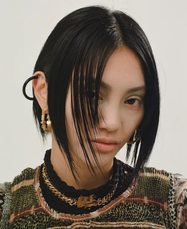 Close-up of a stylish adult female model with short black hair, gold jewelry, and fashionable layered outfit in a studio setting.