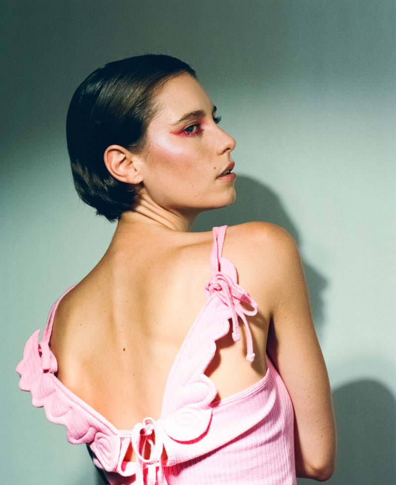 Young woman with short hair, pink eye makeup, and wearing a pink top with ruffles, posing in front of a light blue background.