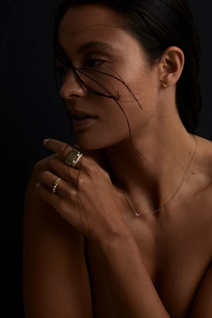 Close-up of a woman with dark hair and neutral makeup wearing jewelry. She has rings on her fingers and a delicate necklace.