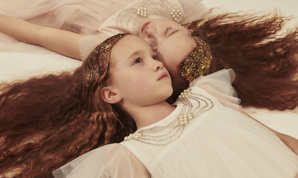 Two young girls lie together with long auburn hair, wearing pearl-adorned dresses. Soft, diffused light, shallow depth of field, studio setting.