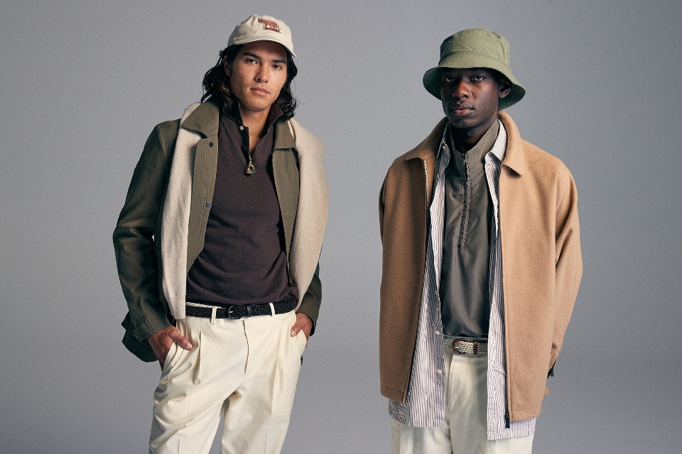 Two men pose in a studio wearing casual attire. One wears a hat, while the other sports a baseball cap. Neutral backdrop.