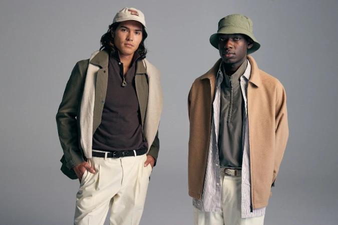 Two men pose in a studio wearing casual attire. One wears a hat, while the other sports a baseball cap. Neutral backdrop.