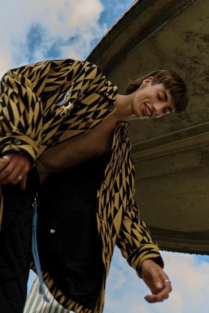 Young adult poses outdoors wearing a patterned jacket and black pants, with a unique mullet hairstyle, ring jewelry, and smiling expression.
