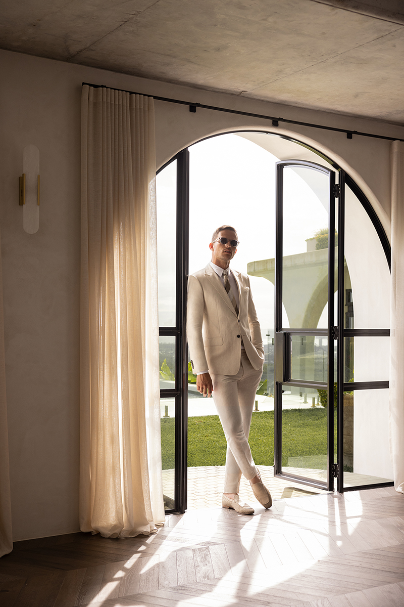 A man in a cream-colored suit stands in an arched doorway leading to a grassy outdoor area, posing for a fashion or lifestyle shoot.