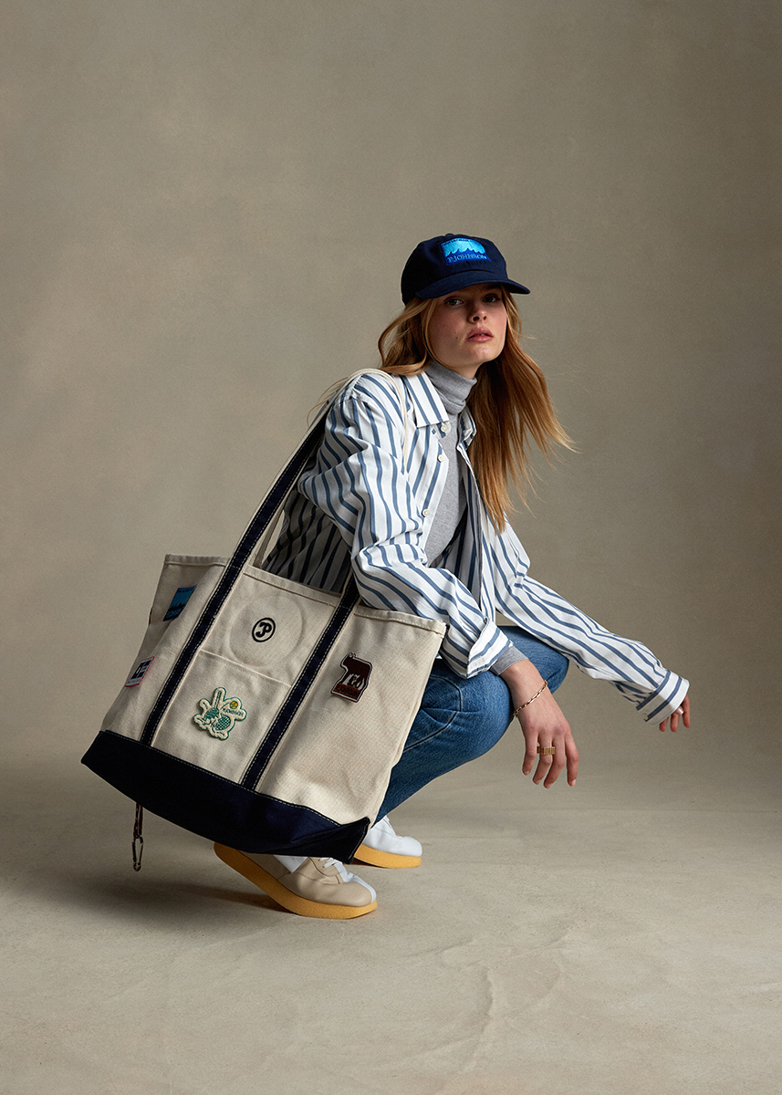 Adult female crouching in a studio wearing a striped shirt, baseball cap, blue jeans, and carrying a tan tote bag with navy trim.