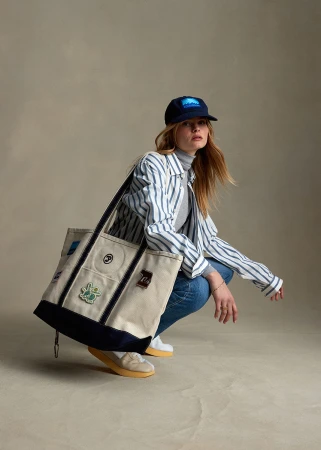 Adult female crouching in a studio wearing a striped shirt, baseball cap, blue jeans, and carrying a tan tote bag with navy trim.