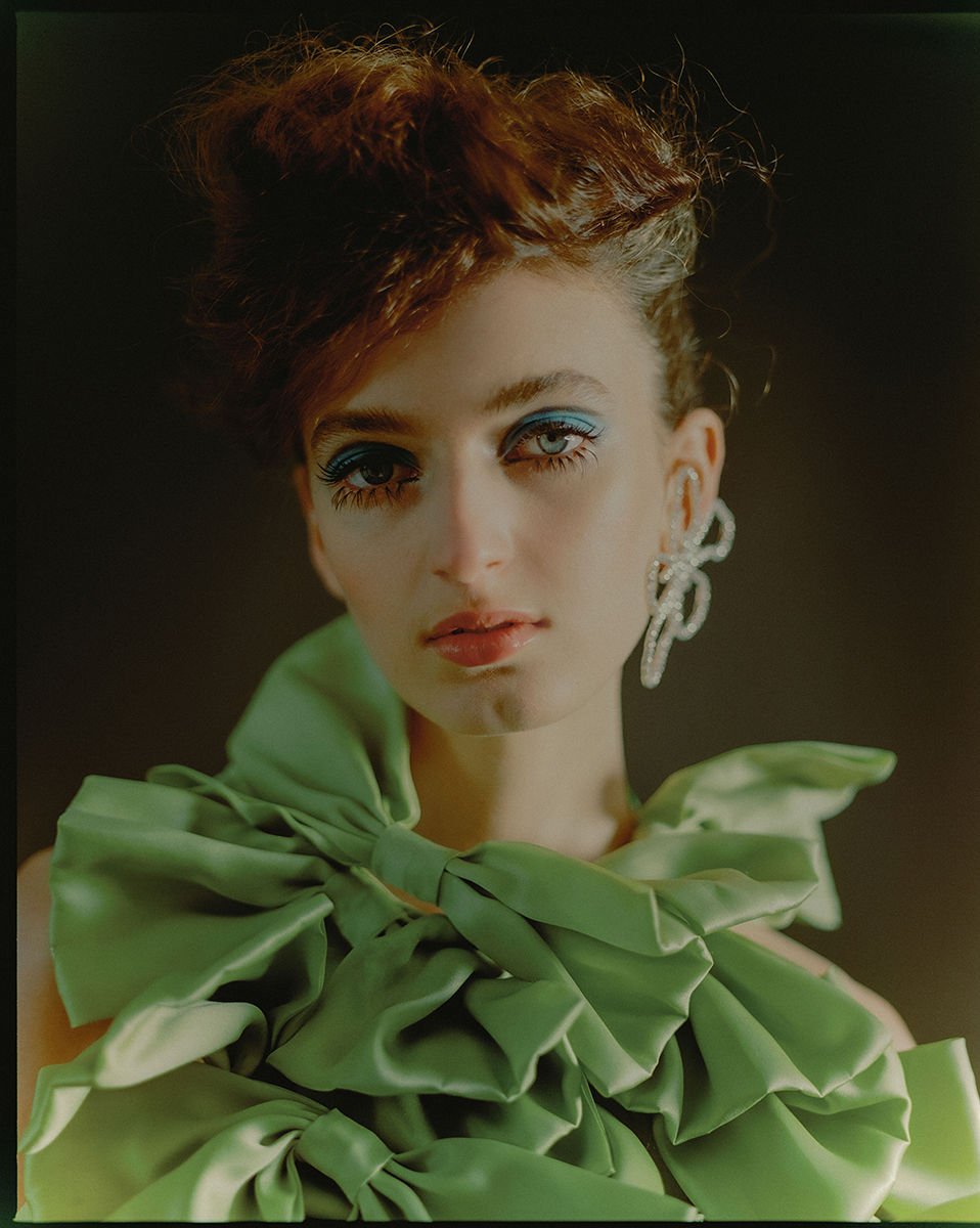 A young woman with red hair and blue eyeshadow poses in a studio, wearing a lime-green gown with large bows and sparkling floral earrings.