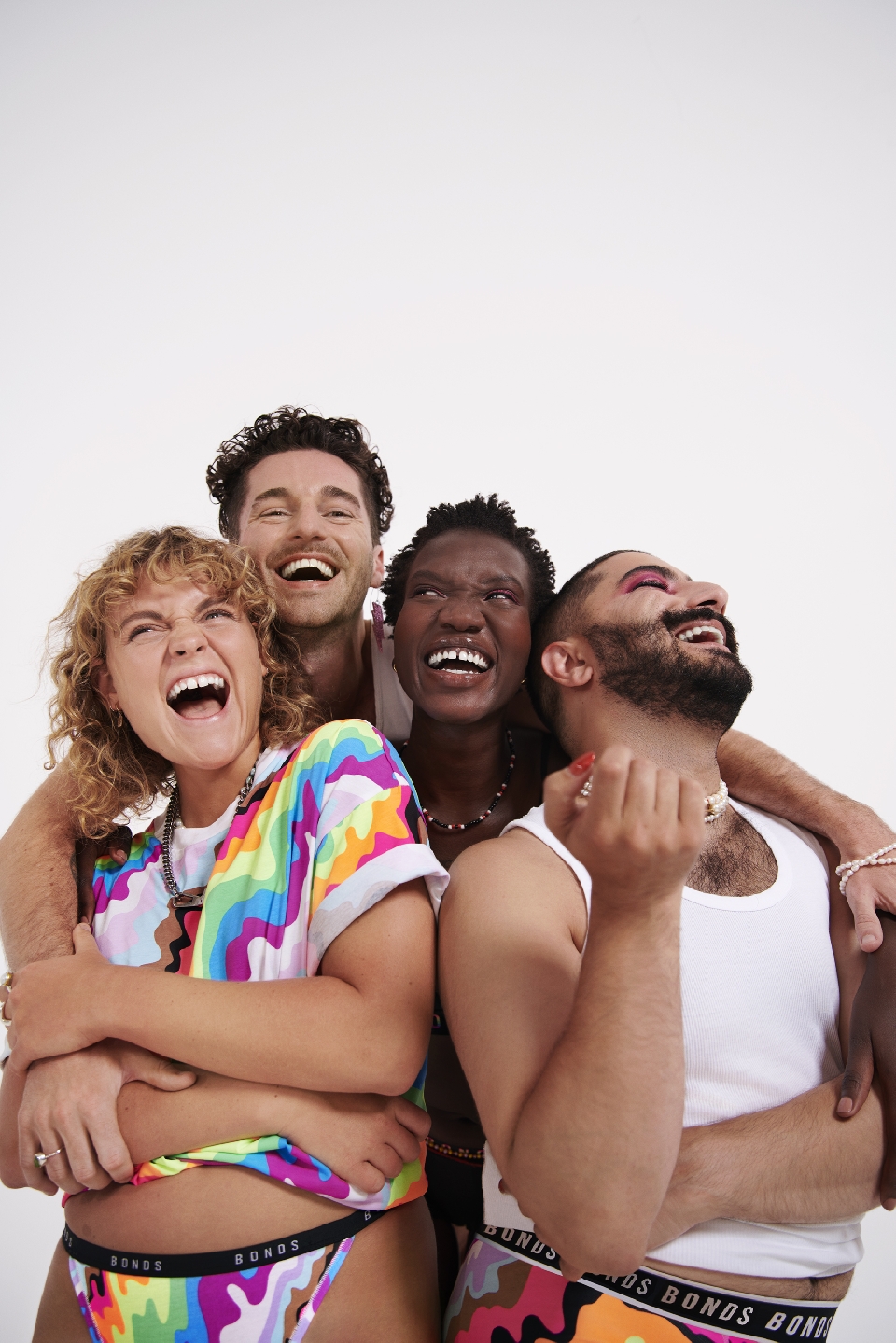 Four people embracing and laughing in a photo studio wearing underwear and rainbow colored tops with a solid white background.