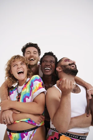 Four people embracing and laughing in a photo studio wearing underwear and rainbow colored tops with a solid white background.