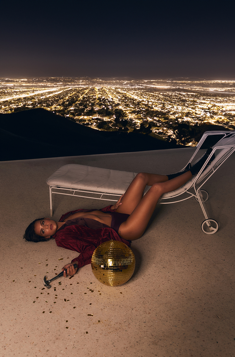 Woman poses in a red outfit on a deck chair with a disco ball, overlooking a sprawling cityscape at night, creating a striking visual.