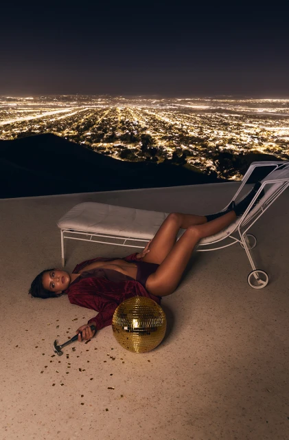 Woman poses in a red outfit on a deck chair with a disco ball, overlooking a sprawling cityscape at night, creating a striking visual.